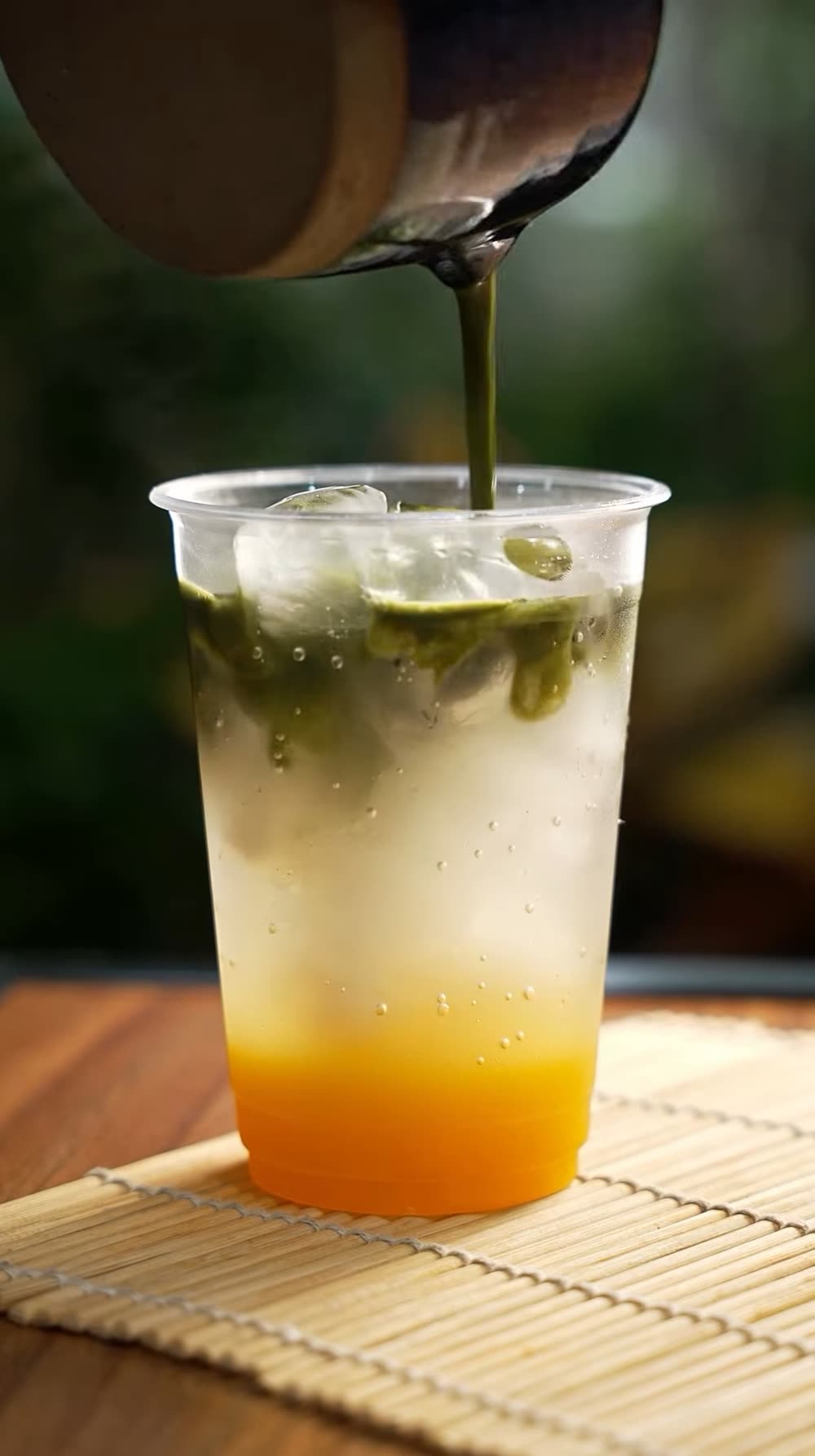 Matcha sendo servido sobre gelo e maracujá num copo, com as plantas do jardim ao fundo